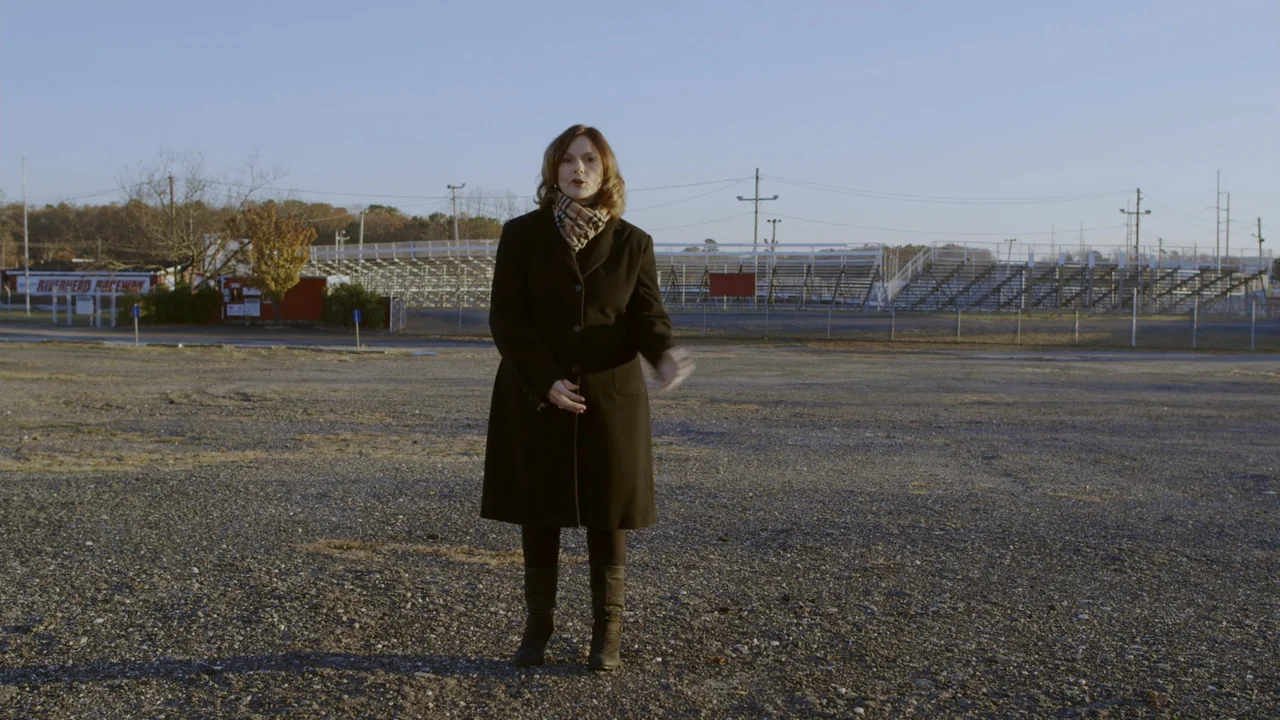 The Last Race (2022) movie still: fullbody — An expansive, gravel-filled parking lot stretches out in front of metal bleachers at dusk. A woman in a…