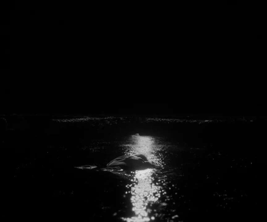 The Lighthouse (2019) movie still: wide — A dark body of water stretches across the frame at night. A bright light reflects intensely on the water…