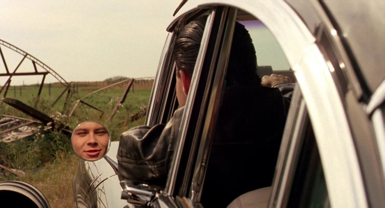 The Reflecting Skin (1990) movie still: medium — A field with a broken metal structure visible through a car window. A man in a leather jacket sits…
