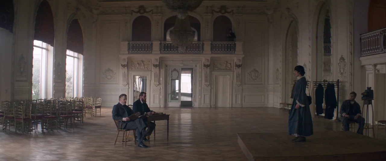 The Dancer movie still: wide — A spacious high-ceilinged ballroom features ornate white wall moldings and polished wooden floors. A woman stands on…