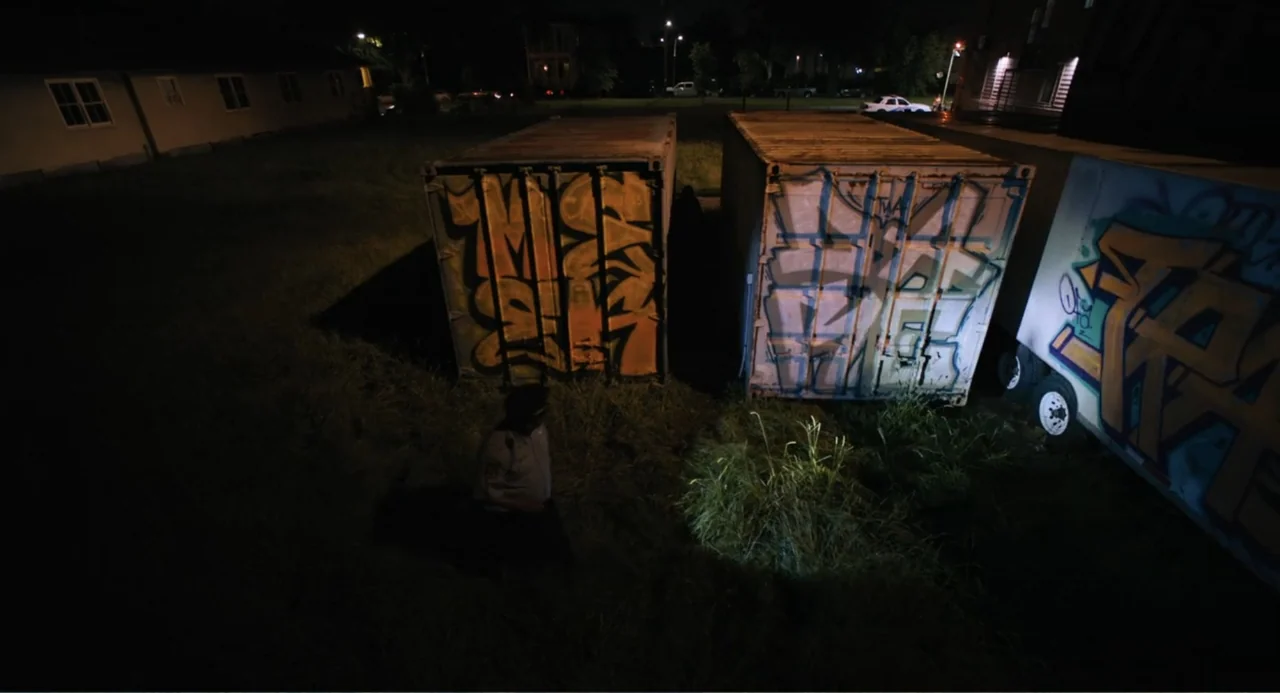 Mona Lisa and the Blood Moon (2022) movie still: wide — A nighttime outdoor scene features two graffiti-covered shipping containers in a…