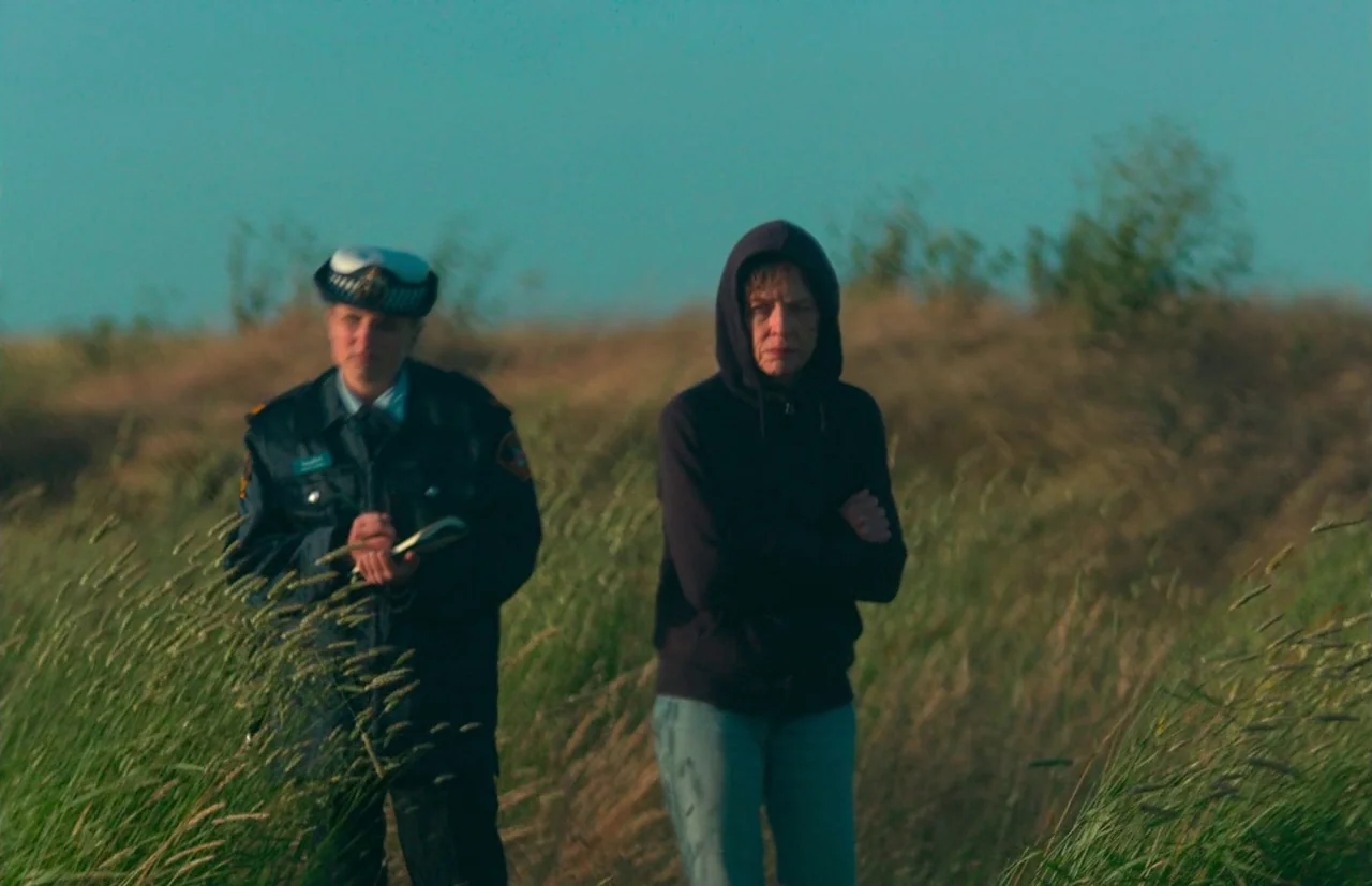 Nitram (2021) movie still: medium — A field of tall grass extends toward the horizon under a clear sky. A woman in a dark hoodie stands beside a…
