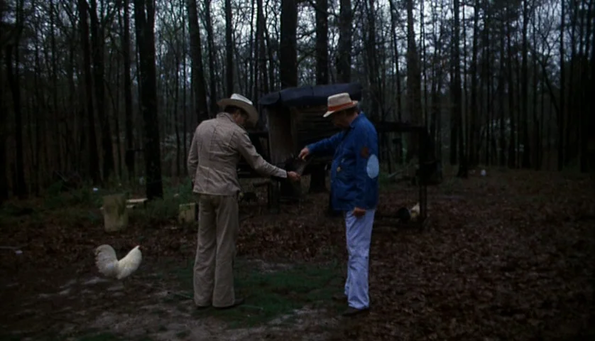 Cockfighter (1974) movie still: fullbody — A dense, leaf-covered wooded area contains a small weathered wooden structure. Two men stand beside a…