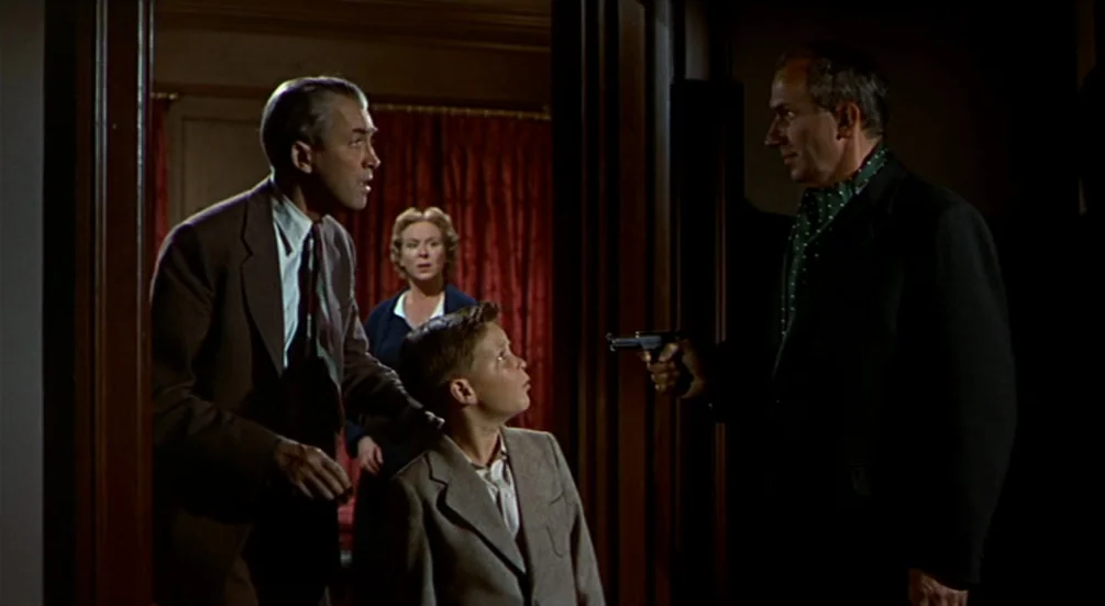 The Man Who Knew Too Much (1956) movie still: medium — A dark, wood-paneled residential space with red curtains visible through a doorway. A man…