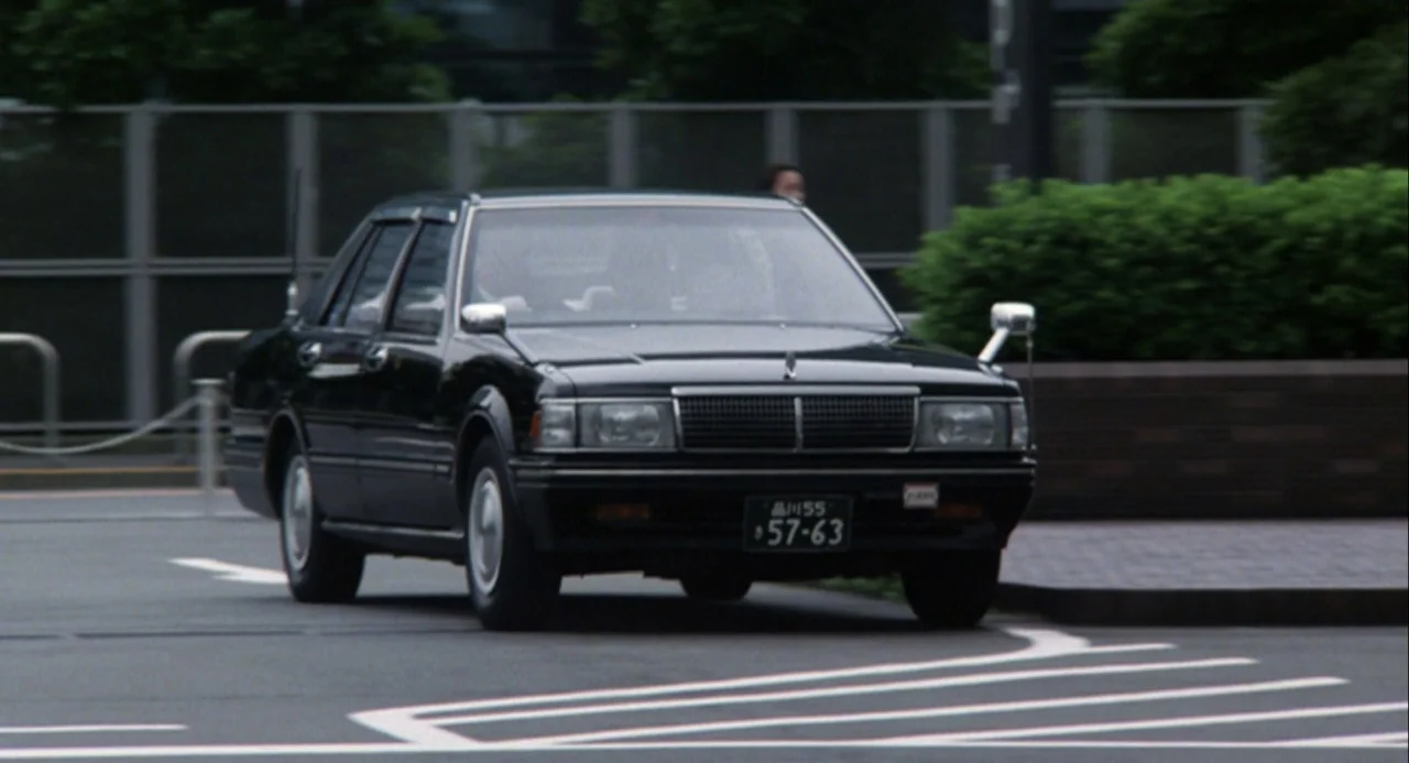 Godzilla and Mothra: The Battle for Earth (1992) movie still: wide — A black sedan turns onto a road surface marked with white lane lines. In the…