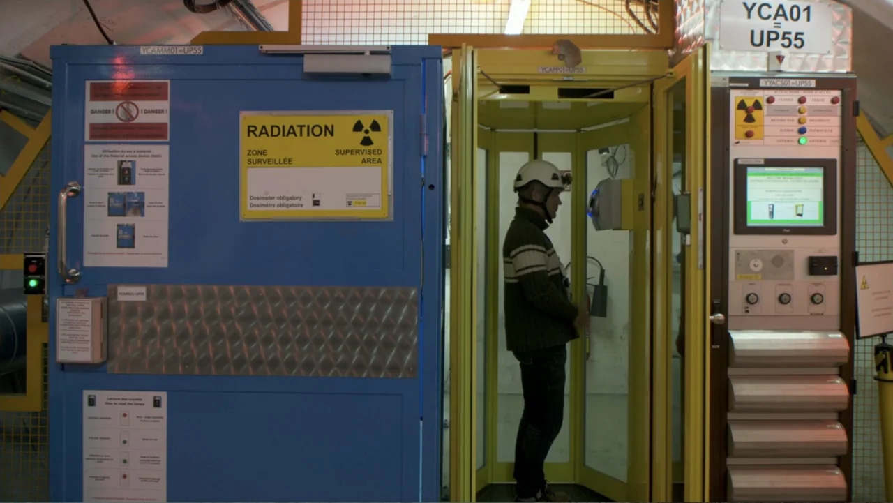 CERN (2013) movie still: medium — An industrial laboratory area features a blue and yellow radiation checkpoint booth. A man wearing a hard hat…