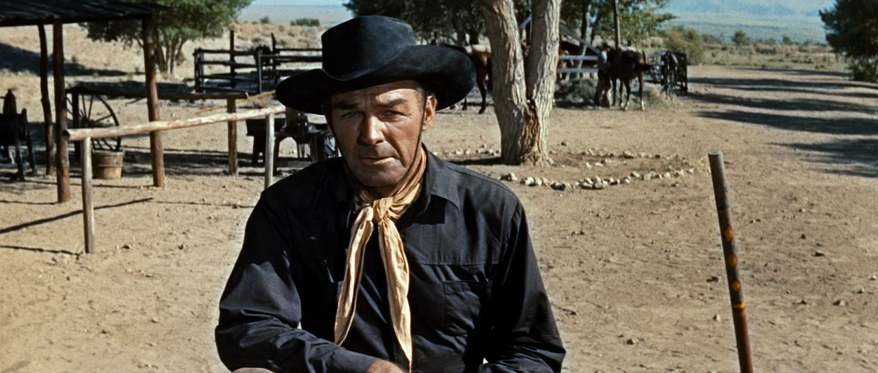 Comanche Station (1960) movie still: medium — A dusty outdoor corral area features wooden posts, sparse trees, and dry ground. A man in a dark hat…