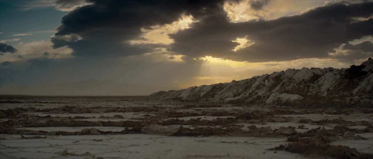 The Neon Demon (2016) movie still: establishing — A vast, cracked salt flat stretches toward a mound of earth under overcast skies. Clouds break to…