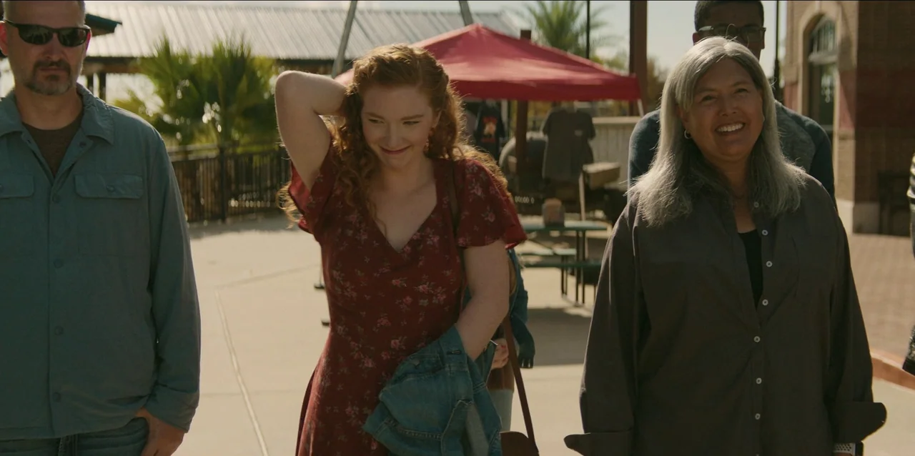The Life of Chuck (2024) movie still: medium — A sunny outdoor plaza features a red tent and tables in the background. Three people walk together…