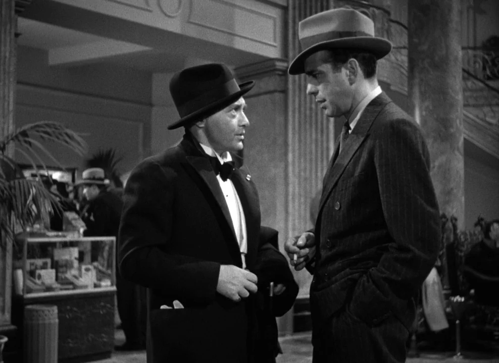 The Maltese Falcon (1941) movie still: medium — A grand hotel lobby features marble columns and a glass display case with merchandise. Two men stand…
