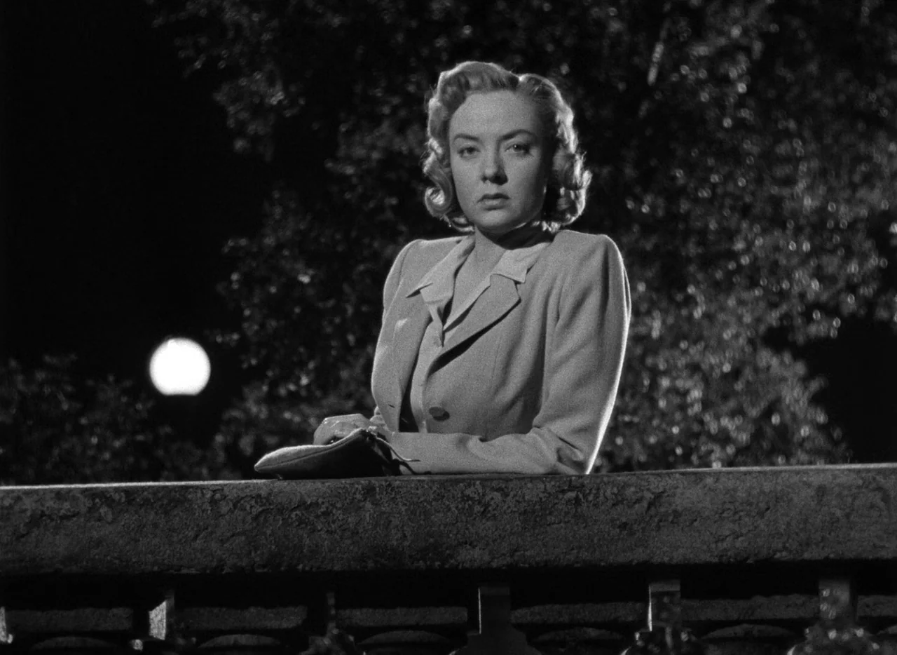 The Set-Up (1949) movie still: medium — A woman stands behind a stone railing at night with dark trees visible behind her. She leans forward with…