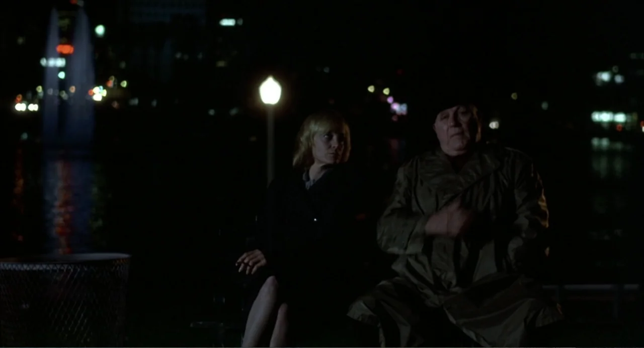 Murphy's Law (2009) movie still: medium — A park at night is illuminated by a single streetlamp over a bench. A man in a trench coat and a woman sit…