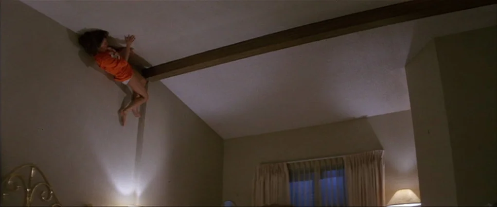 Poltergeist movie still: wide — A hotel room features a standard bed layout with warm bedside lamp lighting. A young girl in an orange shirt crawls…