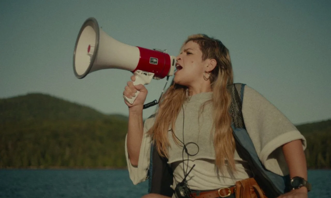 Black Bear (2020) movie still: medium — A person stands on a boat with a large lake and distant hills behind. The subject speaks into a megaphone…