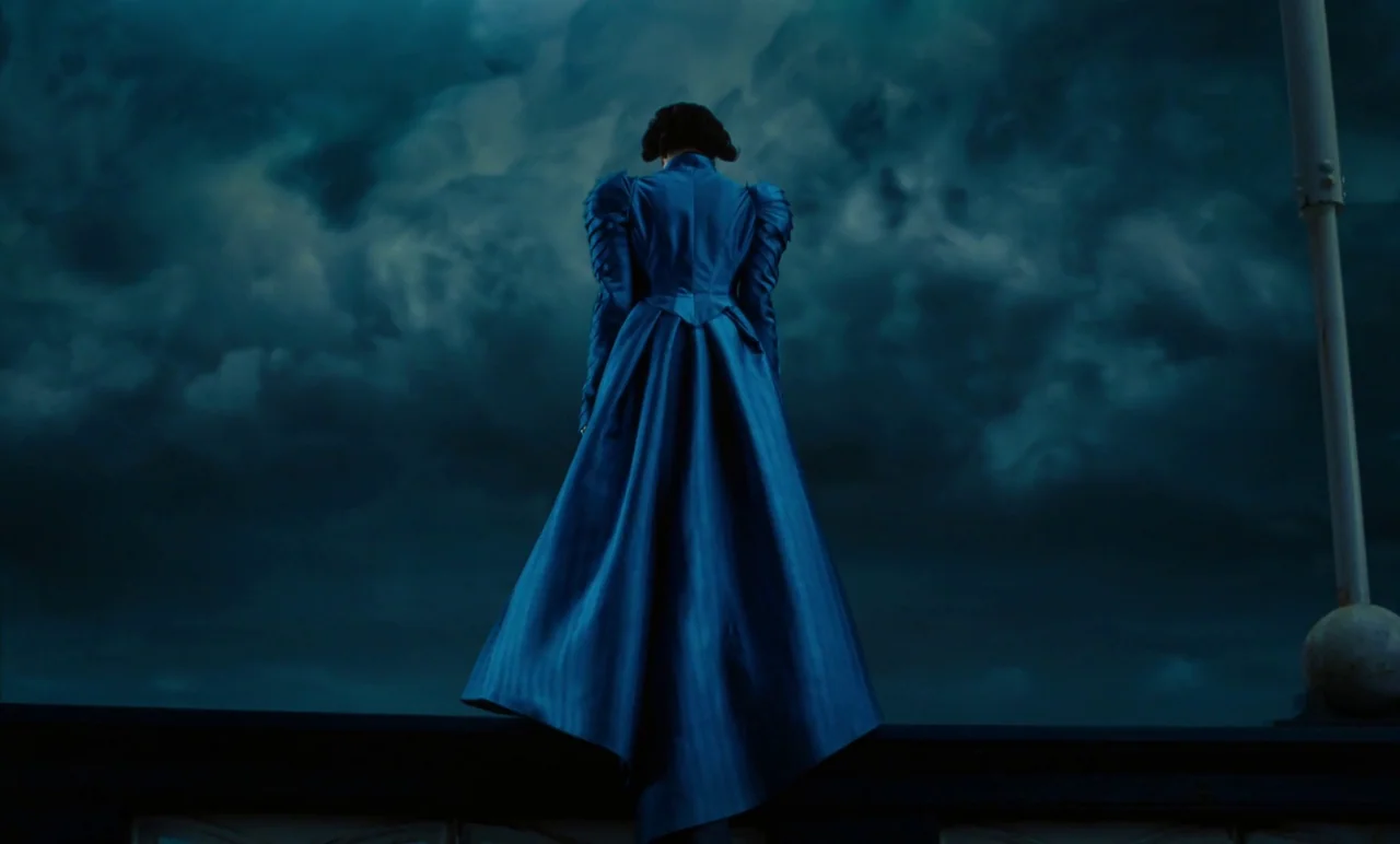 Poor Things (2023) movie still: fullbody — A woman stands atop a building ledge under a vast, dark, turbulent sky. She looks out over the horizon…
