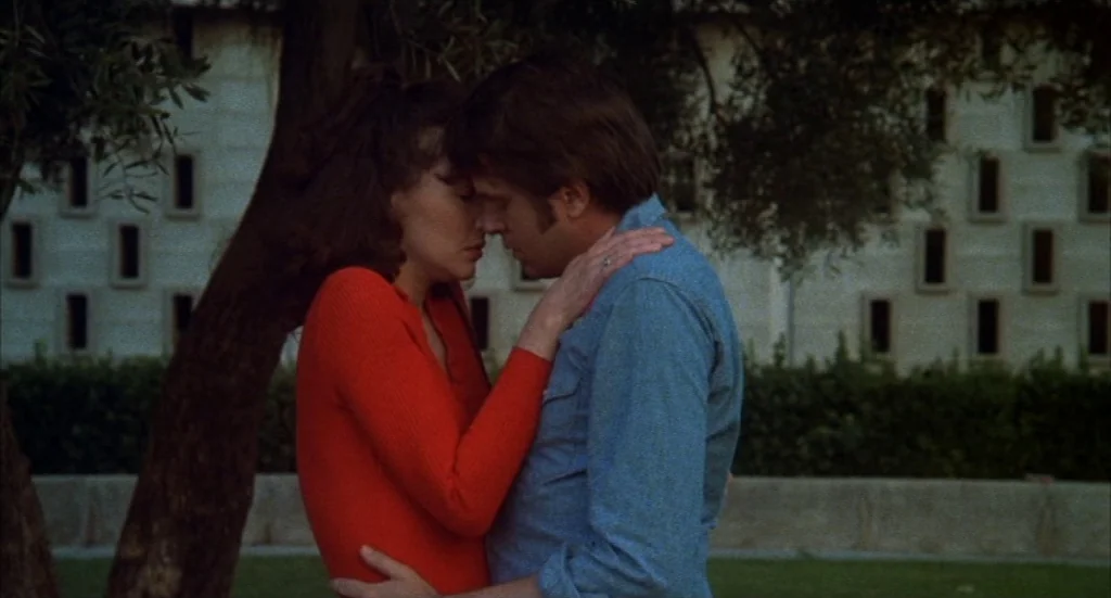 Melvin & Howard (1980) movie still: medium — A man and a woman stand in a park in front of an olive tree. The background features a large stone…