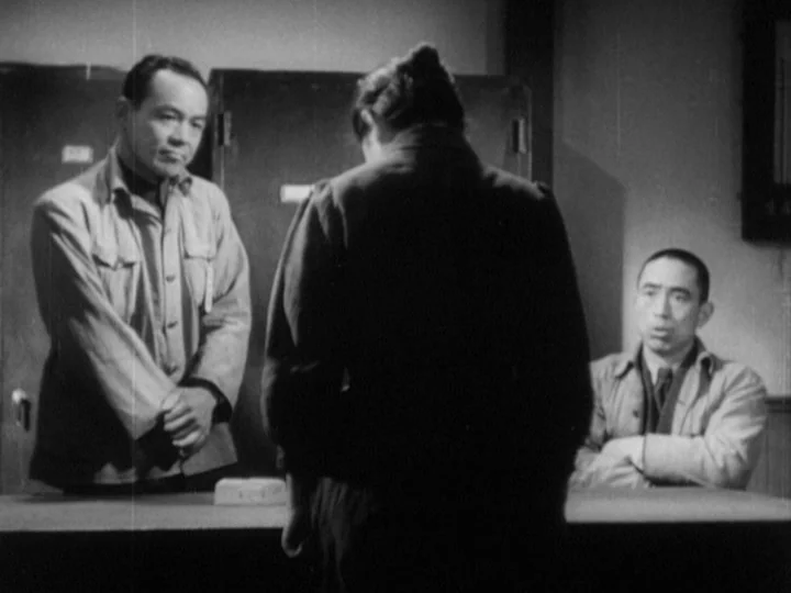 The Most Beautiful (1944) movie still: medium — A spartan room features three people around a central table with dark cabinets behind. A woman…