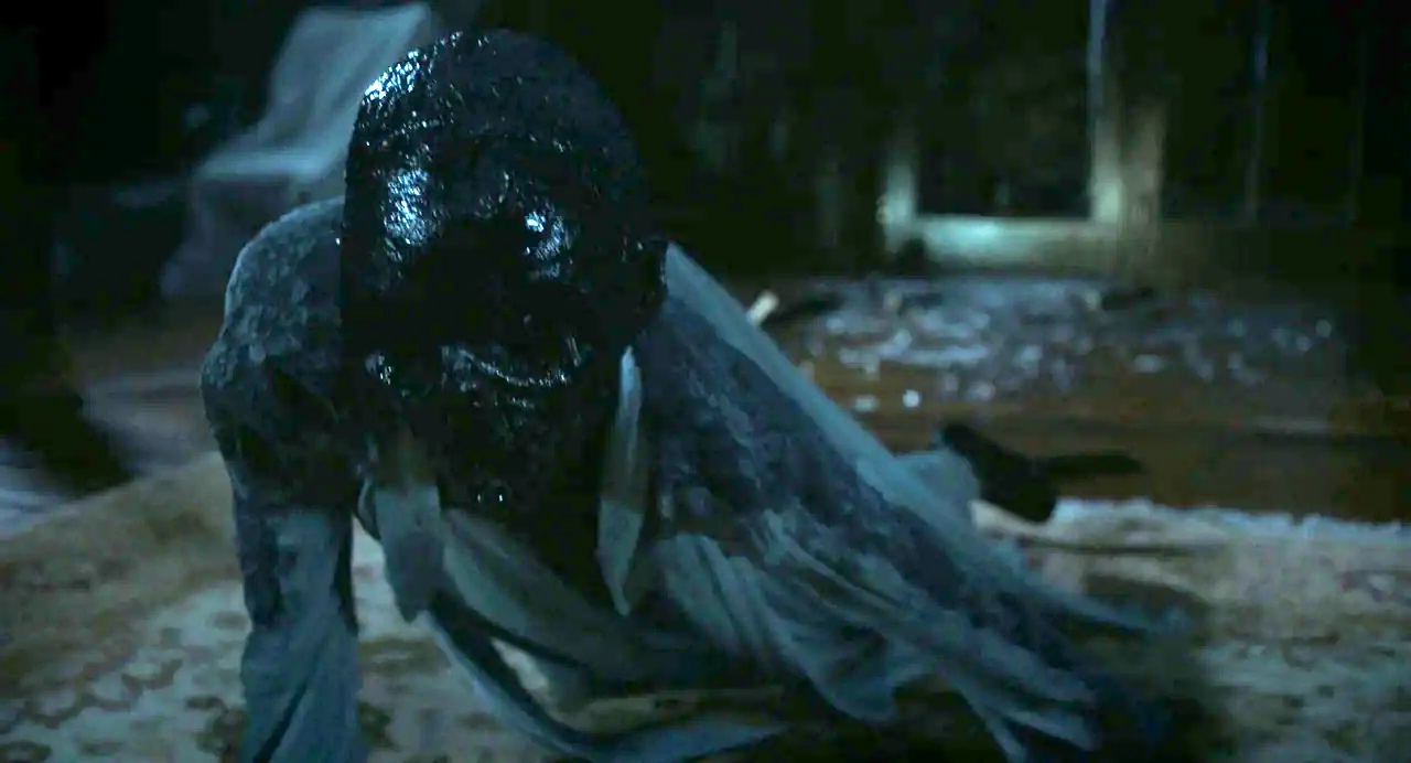 Goodnight Mommy (2022) movie still: closeup — A dark, dimly lit room contains a person crawling on a patterned rug. The figure has a black,…