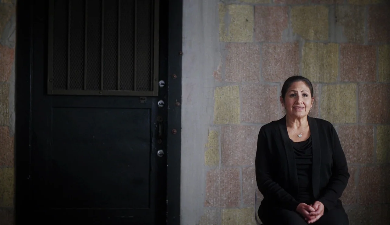 13th (2016) movie still: medium — A woman sits in a chair in front of a heavy black metal door. The backdrop consists of exposed, roughly textured…
