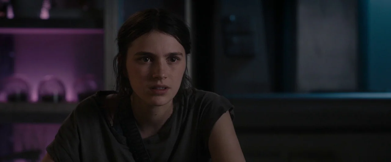 Settlers (2021) movie still: medium — A dark indoor space with subtle purple and teal lighting elements visible in the background. A young woman…