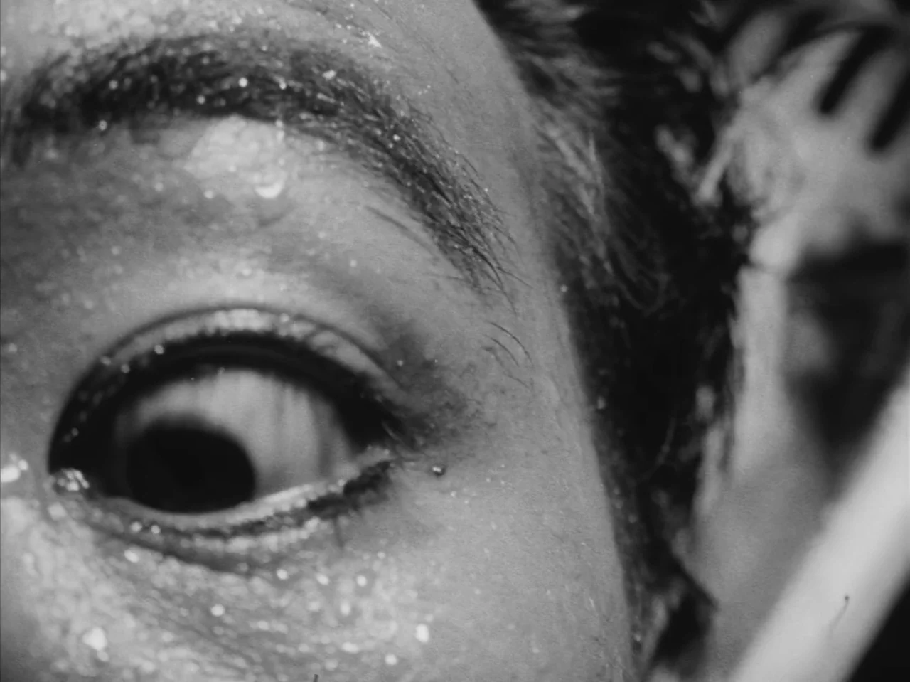 Tetsuo: The Iron Man (1989) movie still: closeup — A close-up shot of a human eye in a tiled space. The skin is covered in water droplets with high…