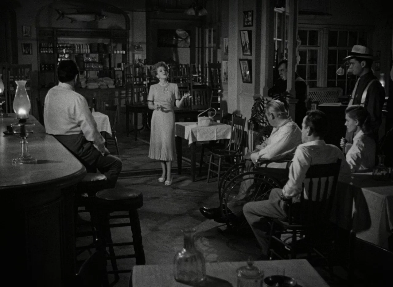 Key Largo (1948) movie still: wide — A dimly lit room contains wooden furniture and several seated individuals in 1940s attire. A woman in a light…