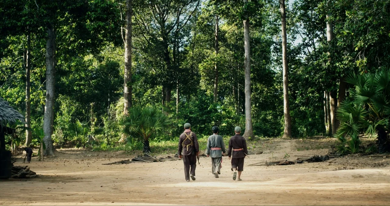 First They Killed My Father (2017) movie still: wide — A rural jungle clearing bordered by tall hardwood trees and lush vegetation. Three men in…