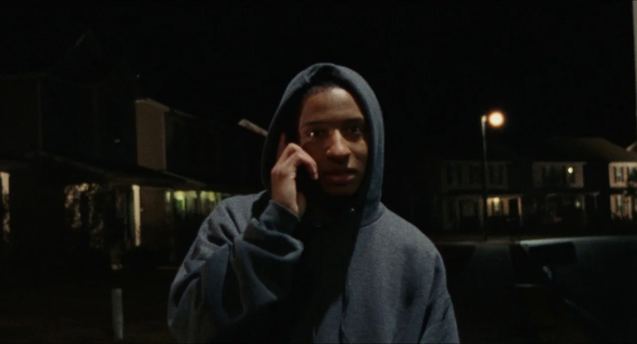 Origin (2023) movie still: medium — A residential street at night with houses visible in the background under darkness. A man in a hooded sweatshirt…