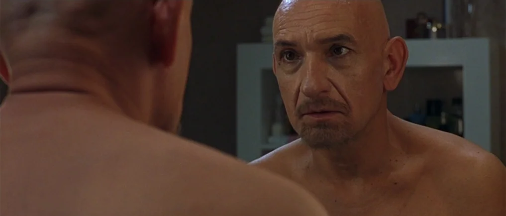 Sexy Beast (2000) movie still: medium — A man stands in front of a mirror in a dimly lit space. The man looks at his own reflection while his bare…