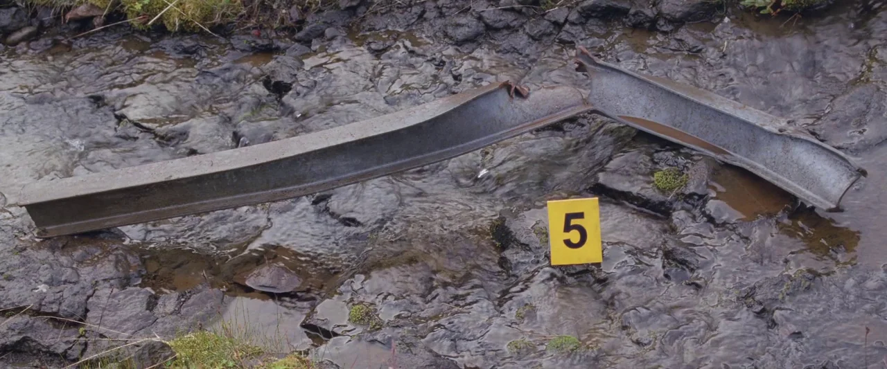 A White, White Day (2019) movie still: closeup — A shallow stream flows over dark, mossy rocks in a rugged outdoor setting. A bent metal beam and a…