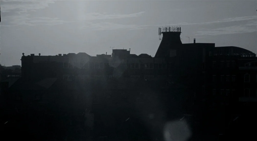 Somers Town (2008) movie still: establishing — An urban skyline of dense buildings appears as a dark silhouette against a bright sky. Several…