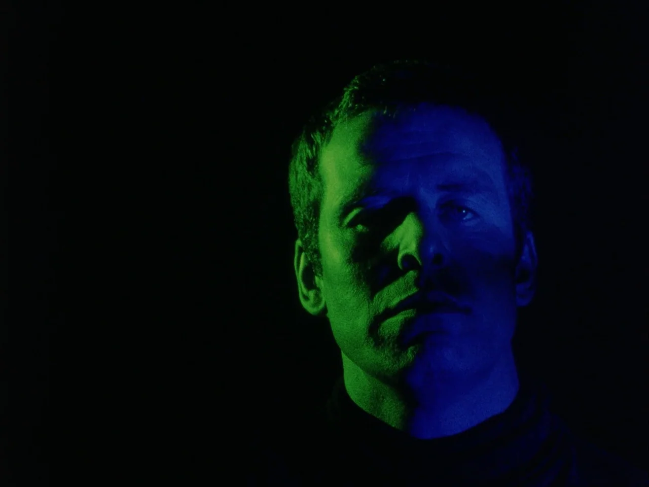 Subconscious Cruelty (2000) movie still: closeup — A man stands against a pitch-black background with no visible architectural details. He faces…