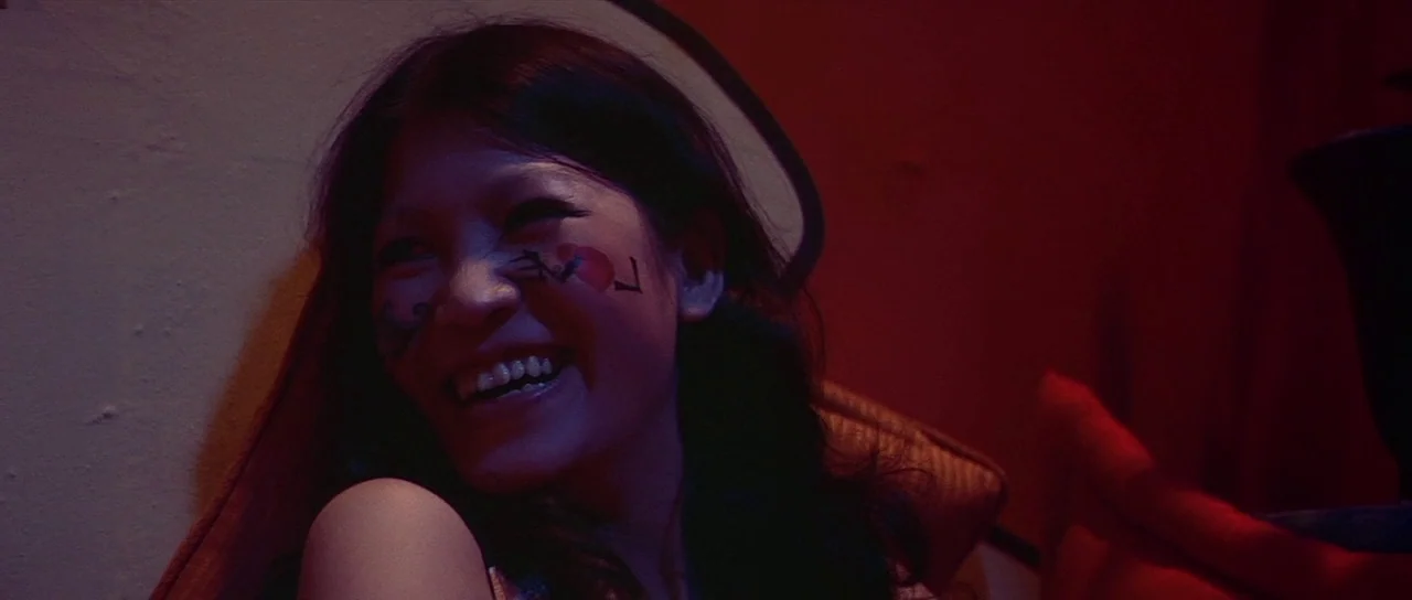 Enter The Dragon (1973) movie still: closeup — A dimly lit room bathed in intense red light. A young woman with writing on her face smiles toward an…