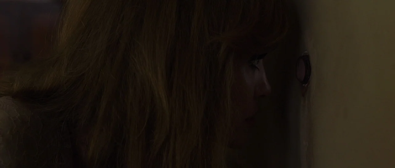 By The Sea (2015) movie still: closeup — A dark, dimly lit corridor features a pale wall with a round metal peephole. A woman presses her eye…