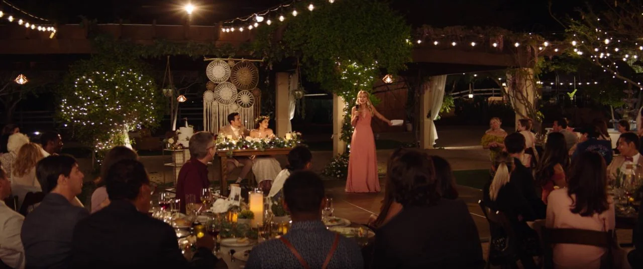 Palm Springs (2020) movie still: wide — An outdoor evening wedding reception features strings of lights draped over a wooden pergola. A woman in a…