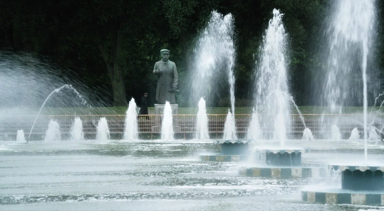 The Death of Stalin (2017) movie still: wide — A series of water fountains erupt from a long rectangular concrete basin in a park. A stone monument…