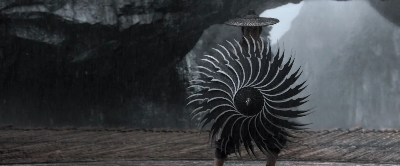 Shadow (2018) movie still: fullbody — A rain-drenched rocky landscape with dark stone cliffs and overcast conditions. A man wearing a conical hat…
