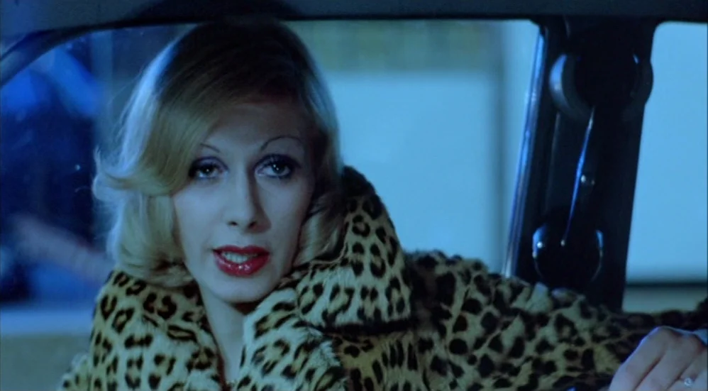 Un Flic (1972) movie still: closeup — A woman sits inside a car at night illuminated by cool exterior ambient light. She wears a leopard-print coat…