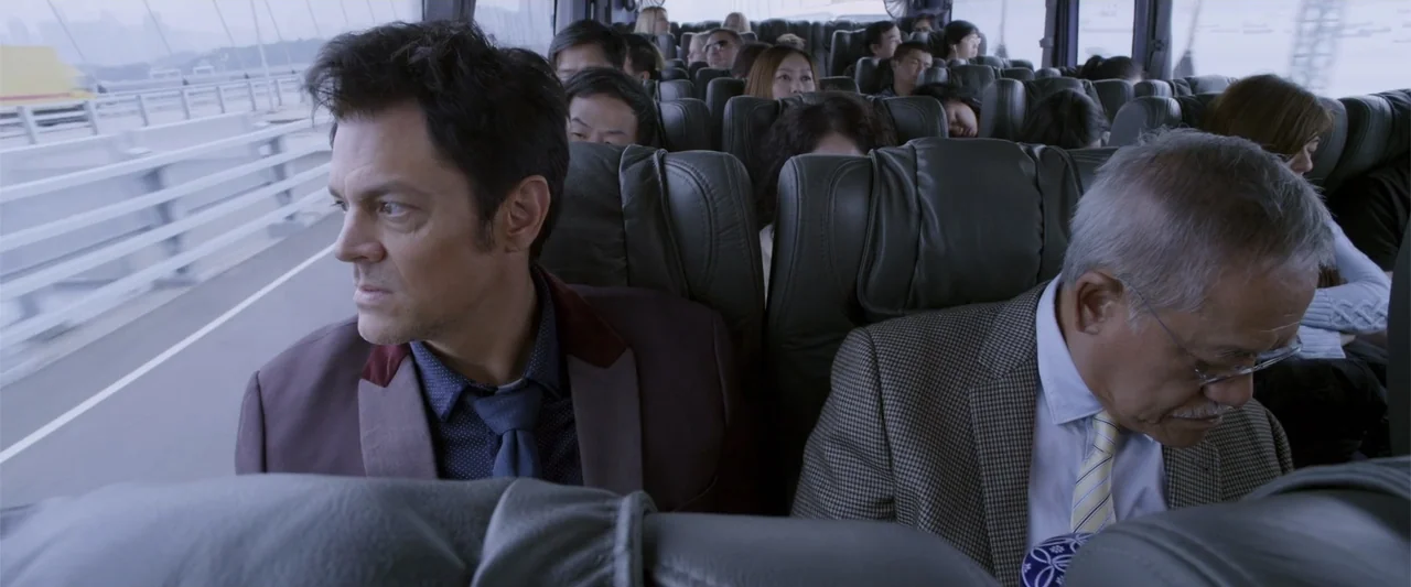 Skiptrace (2016) movie still: medium — The interior of a moving bus with rows of leather seats and blurred window views. Two men sit side by side as…