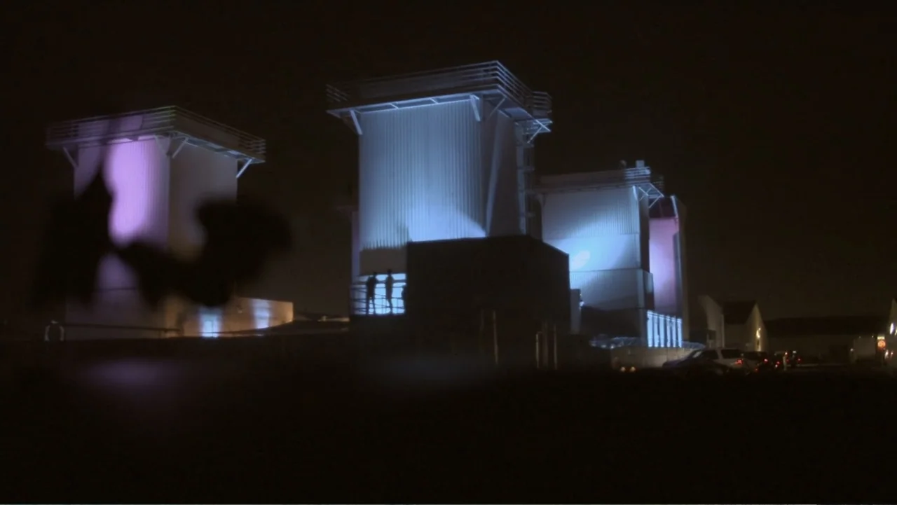Wild Wild Space (2024) movie still: establishing — Large industrial cooling towers stand illuminated against a pitch-black night sky. Silhouetted…
