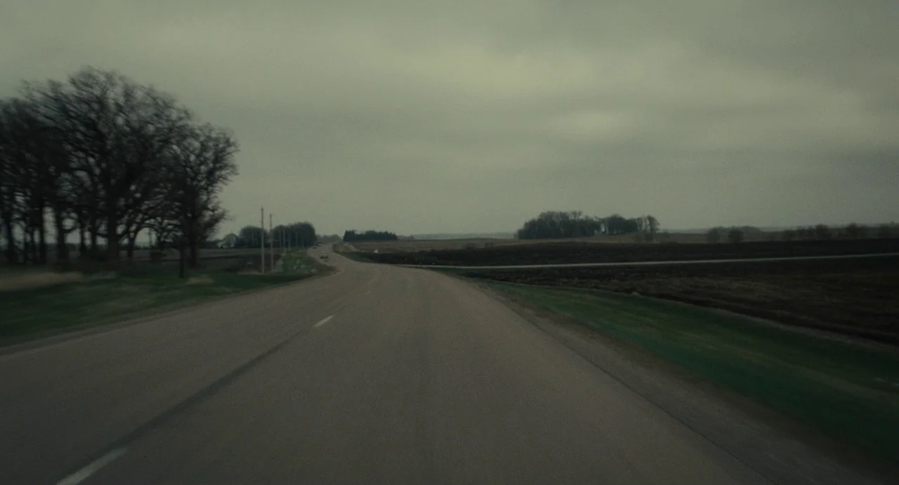 Inside Llewyn Davis (2013) movie still: establishing — An asphalt road curves through flat agricultural fields under a heavy, overcast gray sky.…