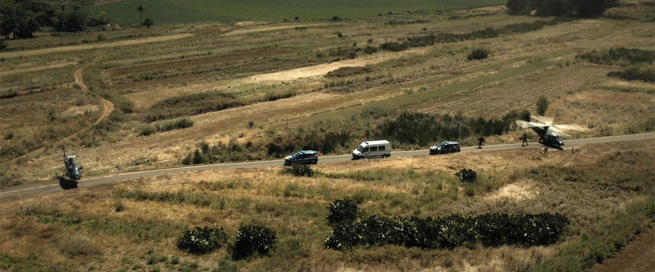 Den of Thieves 2: Pantera (2025) movie still: establishing — A long, paved country road winds through an arid, grass-covered rural landscape. Two…