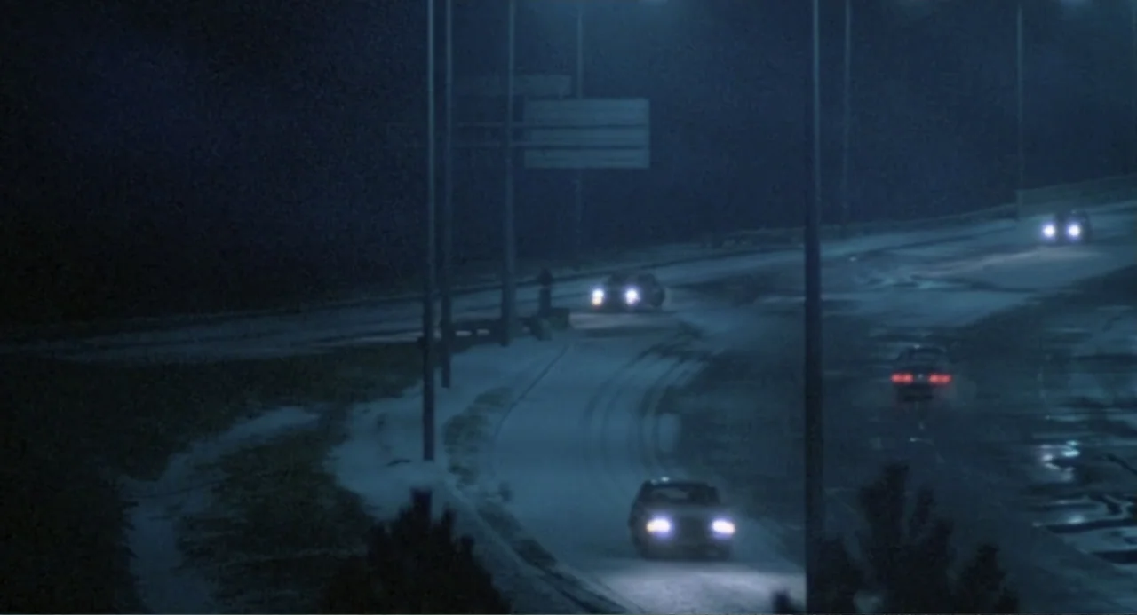 Dead of Winter (2025) movie still: establishing — An elevated view of a multi-lane roadway at night with light snow accumulation. Several cars with…