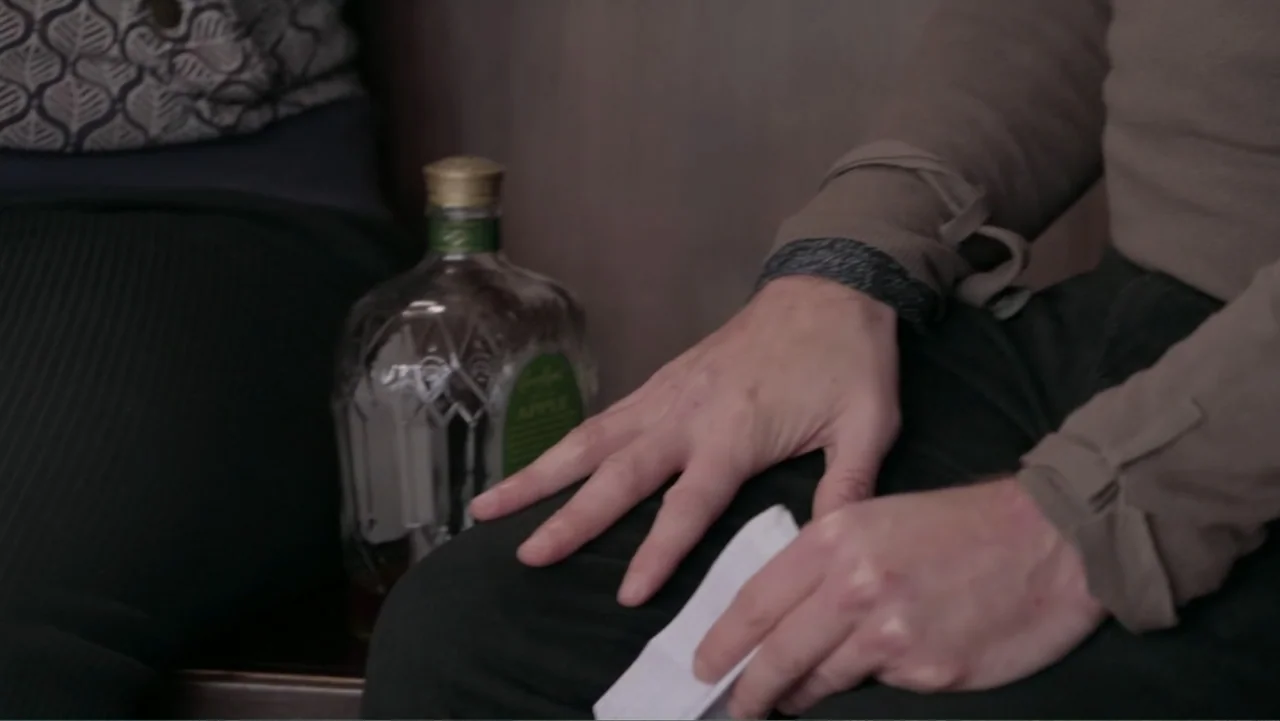 Saturdays Disorders (2022) movie still: closeup — A close view shows a person sitting at a table in a booth. A glass bottle of liquor stands on the…
