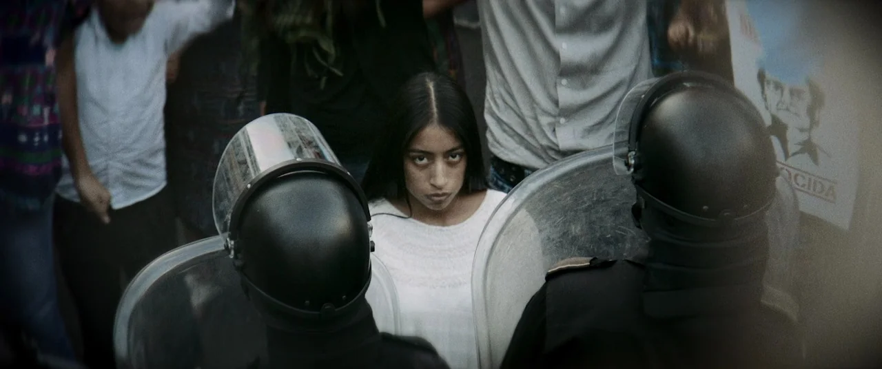 La Llorona (2019) movie still: medium — A dense protest scene features a young woman standing before police riot shields. Officers are positioned in…