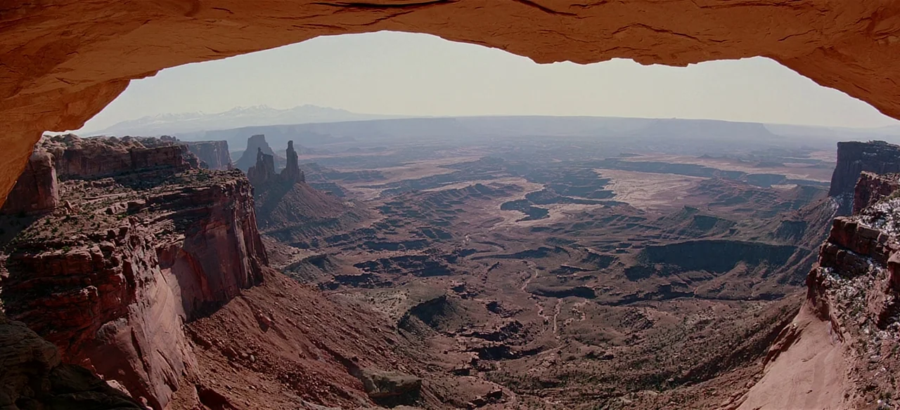 Baraka (1992) movie still: establishing — A vast desert canyon spans the view from inside a large natural rock arch. Deep red sandstone cliffs drop…