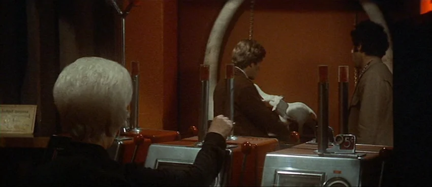 California Split (1974) movie still: medium — A dimly lit arcade room features rows of vintage 5-cent gambling or skill cabinets. Three men stand…
