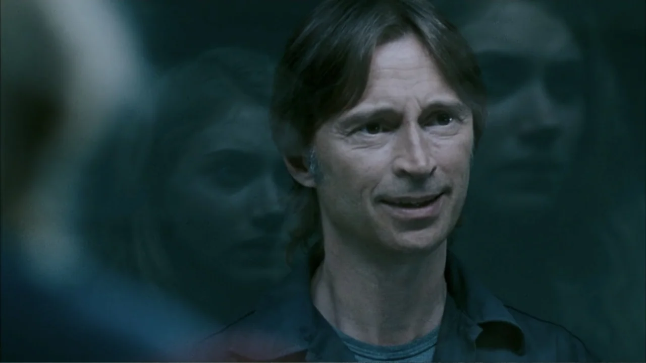 28 Weeks Later (2007) movie still: closeup — A man stands in an indoor space with blurred faces visible in the background. The man faces slightly…