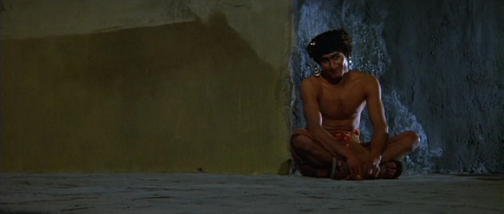 Fellini – Satyricon (1969) movie still: wide — A dark stone corner features a plain, textured surface and a flat floor. A shirtless man sits on the…
