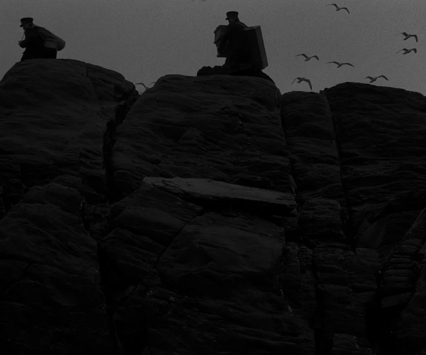 The Lighthouse (2019) movie still: wide — Massive jagged black rock formations rise against a gray, overcast sky. Two figures in dark coats and caps…